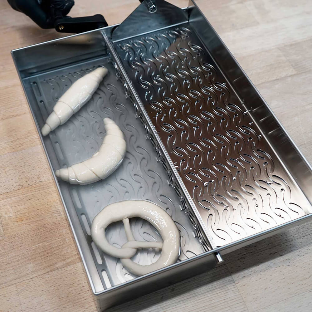 Belaugungsgerät in der Anwendung, es liegen zwei Hörnchen und eine Brezel in der Lauge. Das Gerät steht auf einer Arbeitsplatte.