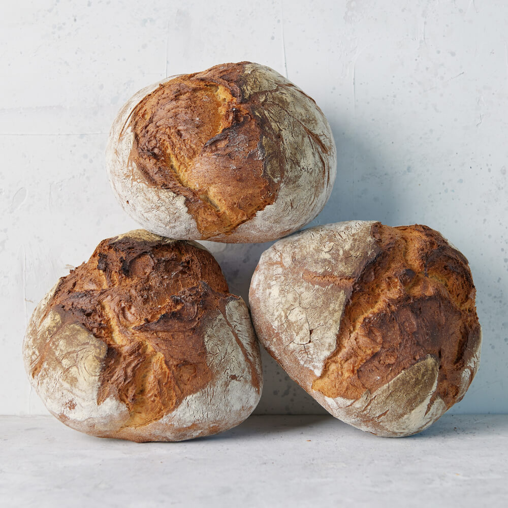 Rezept für Adelindes Schwäbisches Bauernbrot 3 Bauernbrotlaibe die leicht bemehlt sind, aufeinander gestapelt vor einer hellgrauen Wand