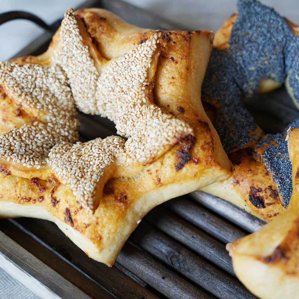 Zwei Sternförmige Brote die in der Mitte ein Loch haben liegen auf einem Gitter. Eines der Brote ist mit Sesam bestreut und das andere mit Mohn.