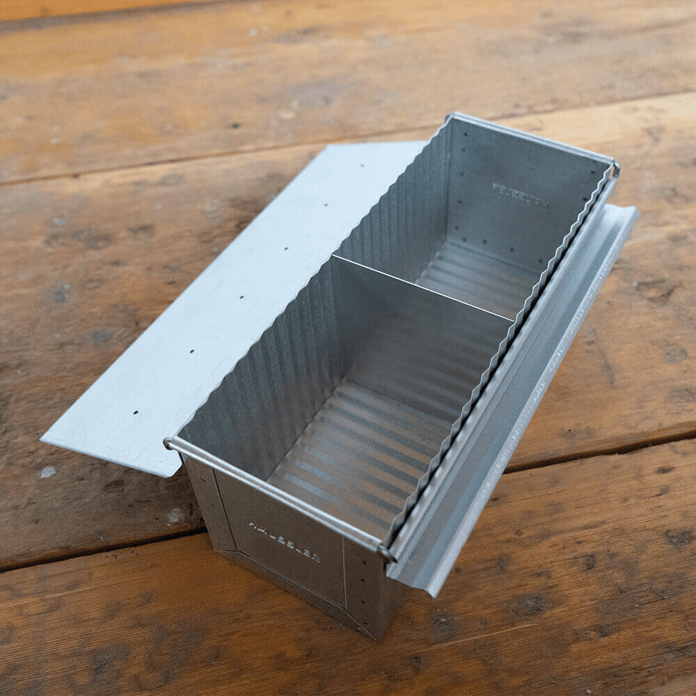 Große Toastbrotform aus aluminiertem Stahl mit geöffnetem Deckel auf einem Holz-Untergrund. Die Abtrennung in der Form ist ersichtlich.