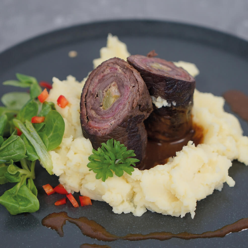 Rezept für Rindsrouladen saftige Rindsrouladen mit Kartoffelpüree und etwas Salat auf einem dunklen Teller angerichtet mit etwas Soße