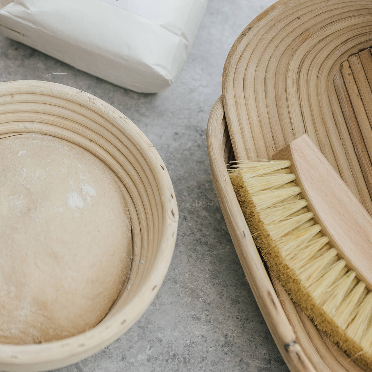 verschiedene_Gärkörbchen_mit_Besen Runde und ovale Gärkörbchen aus Naturmaterial, daneben ein kleiner Besen zum Ausbürsten von Mehlresten.