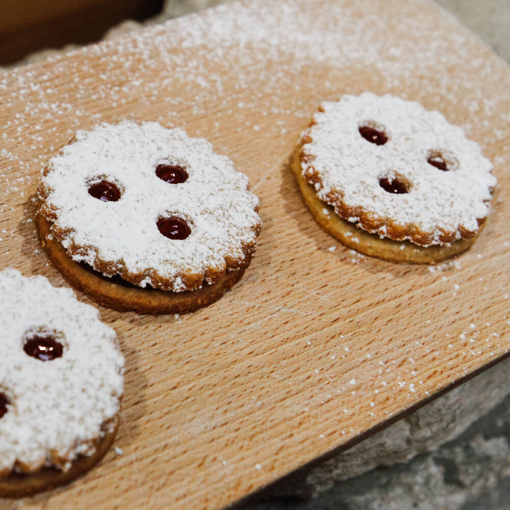 Rezept für Dinkelvollkorn-Spitzbuben 3 Spitzbuben, in runder Form mit welligen Rändern, gefüllt mit roter Marmelade und mit Puderzucker bestreut auf einem Backschieber