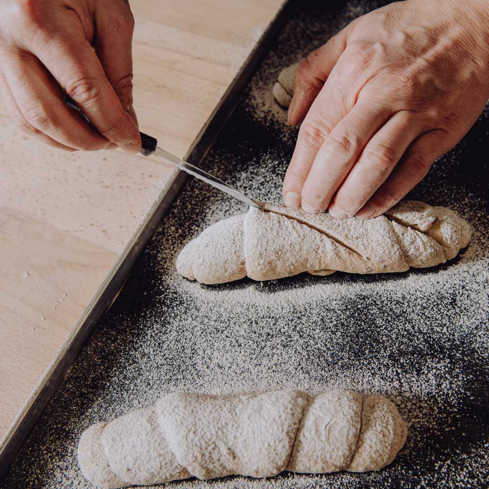 Brötchenteig einschneiden mit einem Brötchenritzmesser Nahaufnahme von Händen, die mit einem Brötchenritzmesser einen Teigling einschneiden, der mit Mehl bestäubt ist