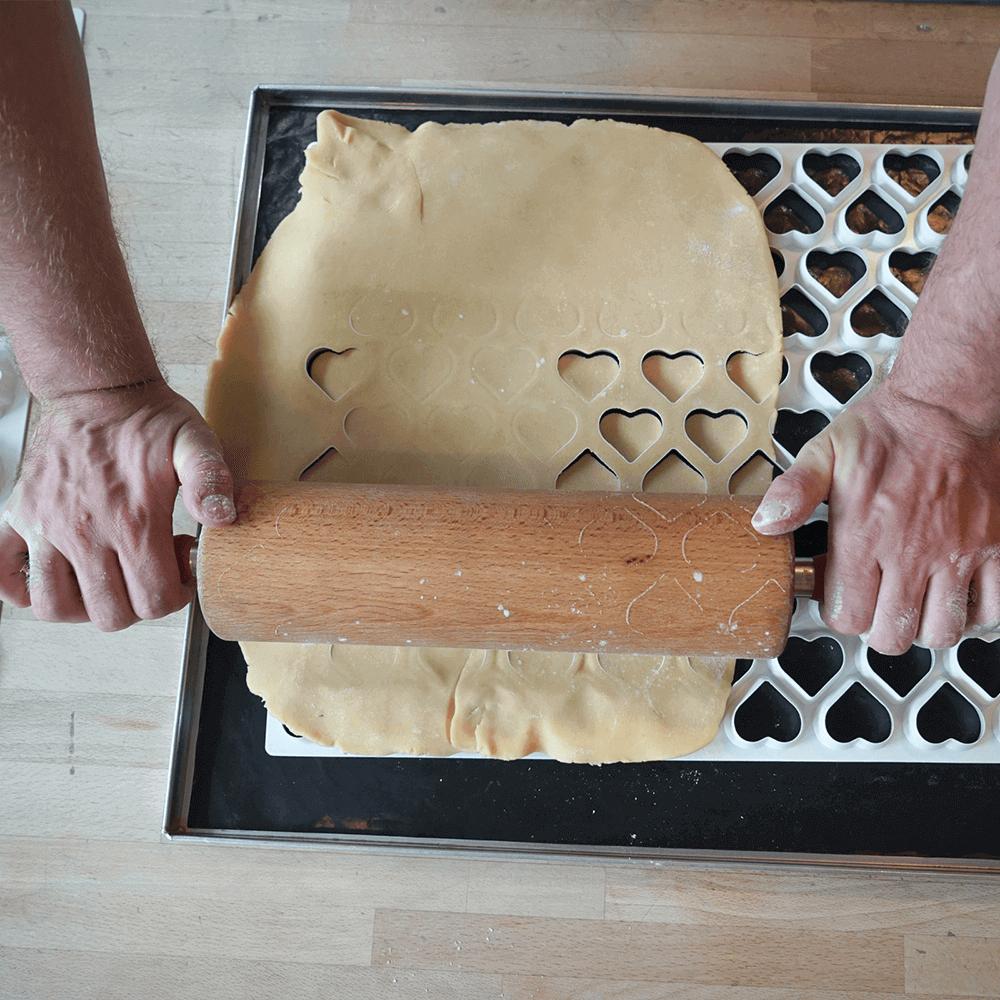 Ausstechmatten für die Plätzchenproduktion Die Ausstechmatte mit dem Muster Herzen wird angewandt. Zwei Hände rollen den Plätzchenteig über die Matte und die ausgestochenen Herzen fallen auf das Backblech.
