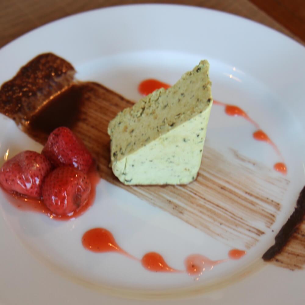 Rezept für Kürbiskern-Parfait mit Vanilleerdbeeren-Ragout Selbstgemachtes Kürbiskernparfait in ein Dreieck geschnitten auf einem weißen Teller mit einem Streifen dunkler Schokolade, Erdbeersoße in Tropfenform und ein wenig Erdbeer-Vanilleragout