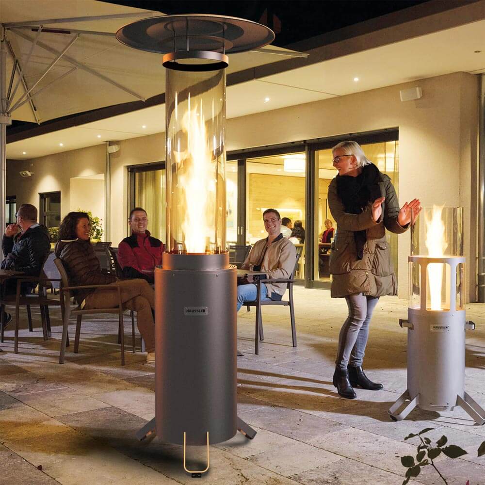 Terrassenstrahler mit angezündetem Feuer auf einer Terrasse. Im Hintergrund sitzen mehrere Personen.