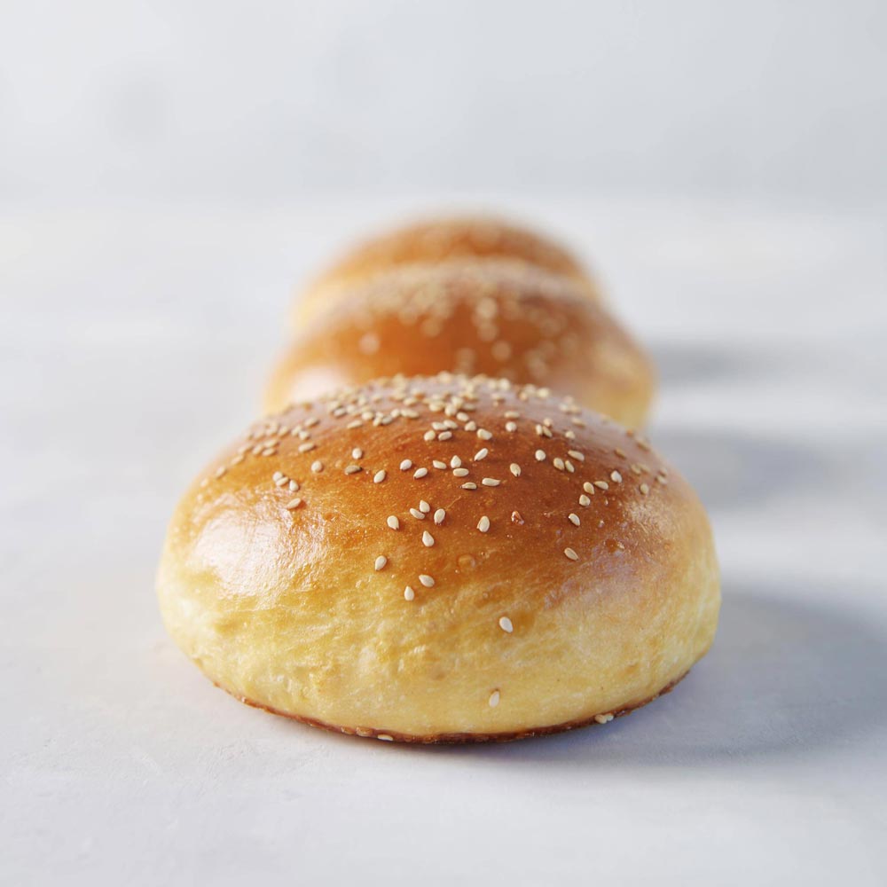 Drei, mit Sesam bestreute, Burger Bun Brötchen liegen hintereinander.