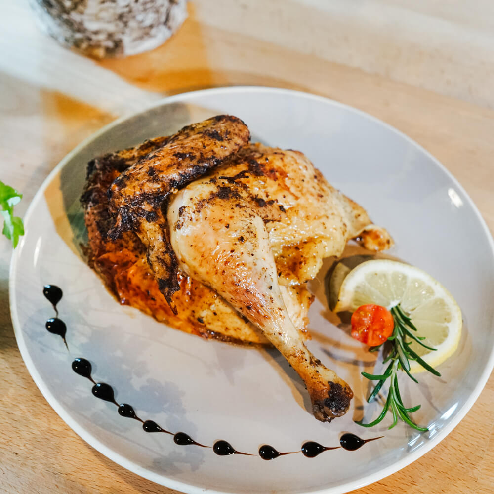 halbes Hähnchen auf einem weißen Teller mit Zitronenscheibe, frischen Kräutern und einer Verzierung aus Balsamico serviert 