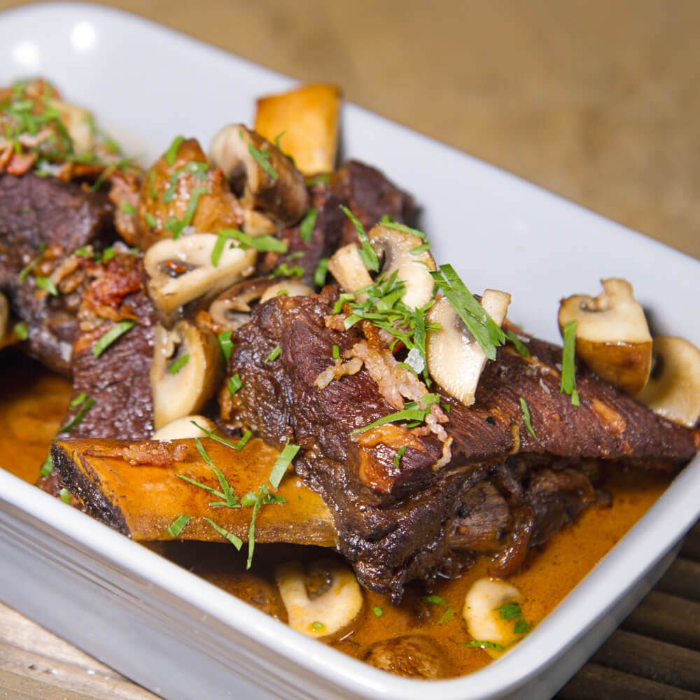 geschmorte Rinderrippen in einer weißen Auflaufform mit braunen Champignons, einer braunen Soße und ein paar Kräutern bestreut