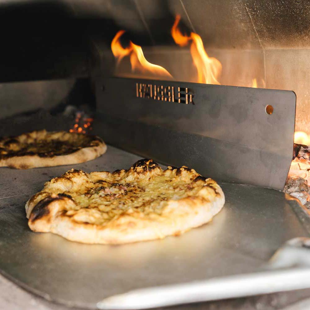 Prallblech für den Holzbackofen Dinnete wird mit Hilfe eines Backschiebers aus dem Ofen geholt. Im Hintergrund ist das Prallblech zu sehen