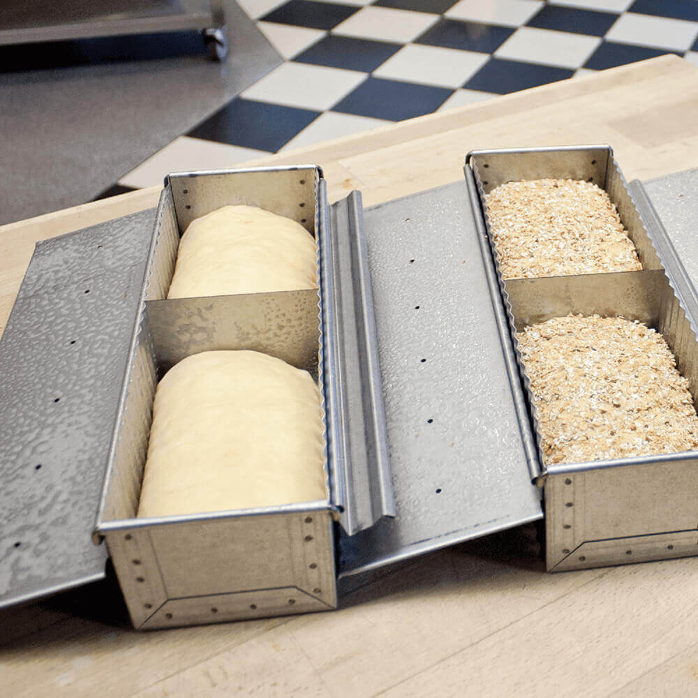 Zwei mittlere Toastbrotformen aus aluminiertem Stahl mit geöffnetem Deckel auf einer Holz-Arbeitsplatte. In beiden Formen befinden sich aufgegangene Toastbrot-Teiglinge, die Teiglinge in der rechten Toastbrotform sind mit Saaten bedeckt.