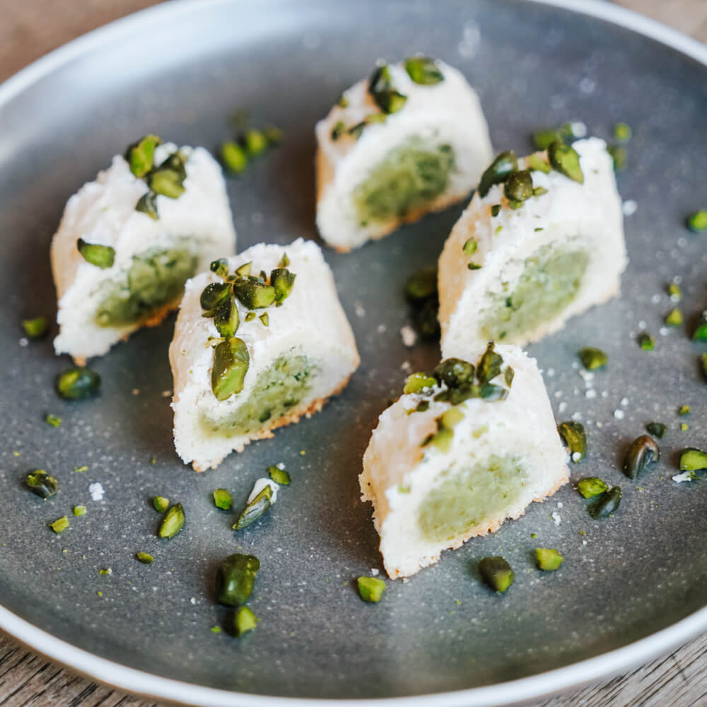 Rezept für Pistazien Röllchen Mürbeteig Rollen gefüllt mit einer Pistazien-Marzipan Füllung in Stücke geschnitten und mit ein paar Pistazien auf dem grauen Teller dekoriert