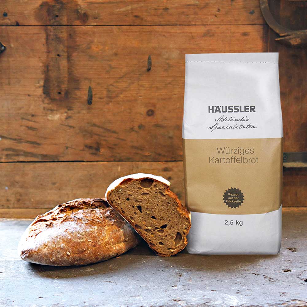 Würziges Kartoffelbrotmehl Würziges Kartoffelbrotmehl in einer 2,5 kg Packung neben einem frisch gebackenem, aufgeschnittenen Kartoffelbrot in einer Küche.