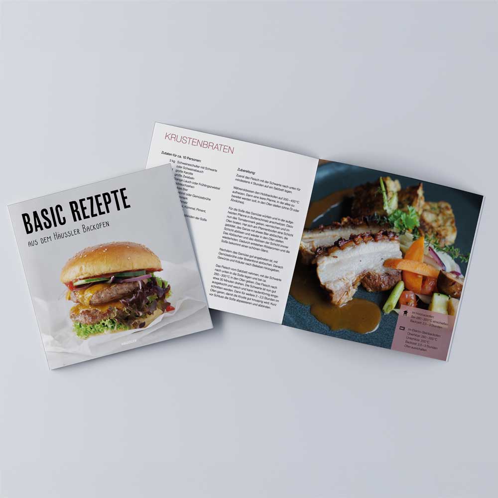 Häussler-Buch: Basic Rezepte aus den Häussler Holzbacköfen Das Häussler-Buch "Basic-Rezepte aus dem Häussler Backofen" liegt aufgeschlagen auf einem grauen Untergrund. Die aufgeschlagene Seite zeigt ein Krustenbraten-Rezept. Daneben liegt ein geschlossenes Buch mit dem Titelbild auf dem ein Burger zu sehen ist.