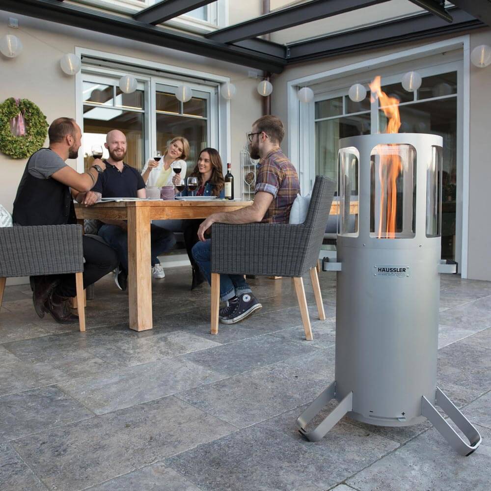 Ambientelicht bei angezündetem Feuer auf einer Terrasse. Im Hintergrund sitzen mehrere Personen und stoßen mit einem Glas Wein an.