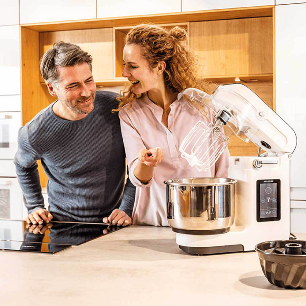 Ein Mann und eine Frau stehen in einer Küche und betrachten die NOVA Teigknetmaschine, welche geöffnet mit dem Schneebeseneinsatz und aufgeschlagenem Eischnee auf einer Küchenarbeitsplatte steht.