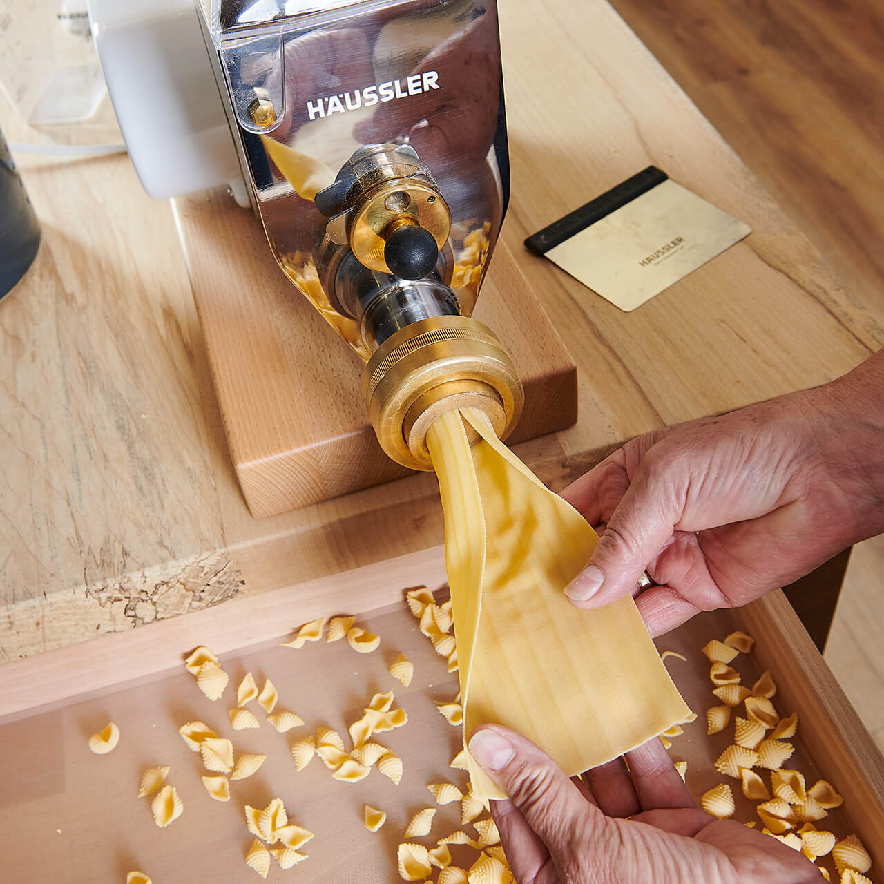 Nudelmaschine_mit_Teig Aus einer Nudelmaschine kommt Maultaschenteig, der von zwei Händen gestützt wird.