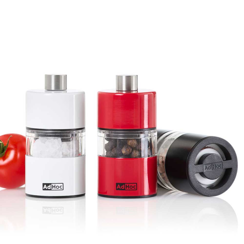 Auf der linken Seite liegt eine Tomate und rechts davon steht eine weiße Mühle mit dem Inhalt Salz: In der Mitte die gleiche Mühle in rot mit Pfeffer, auf der rechten Seite liegt eine Mühle in schwarz mit Pfeffer