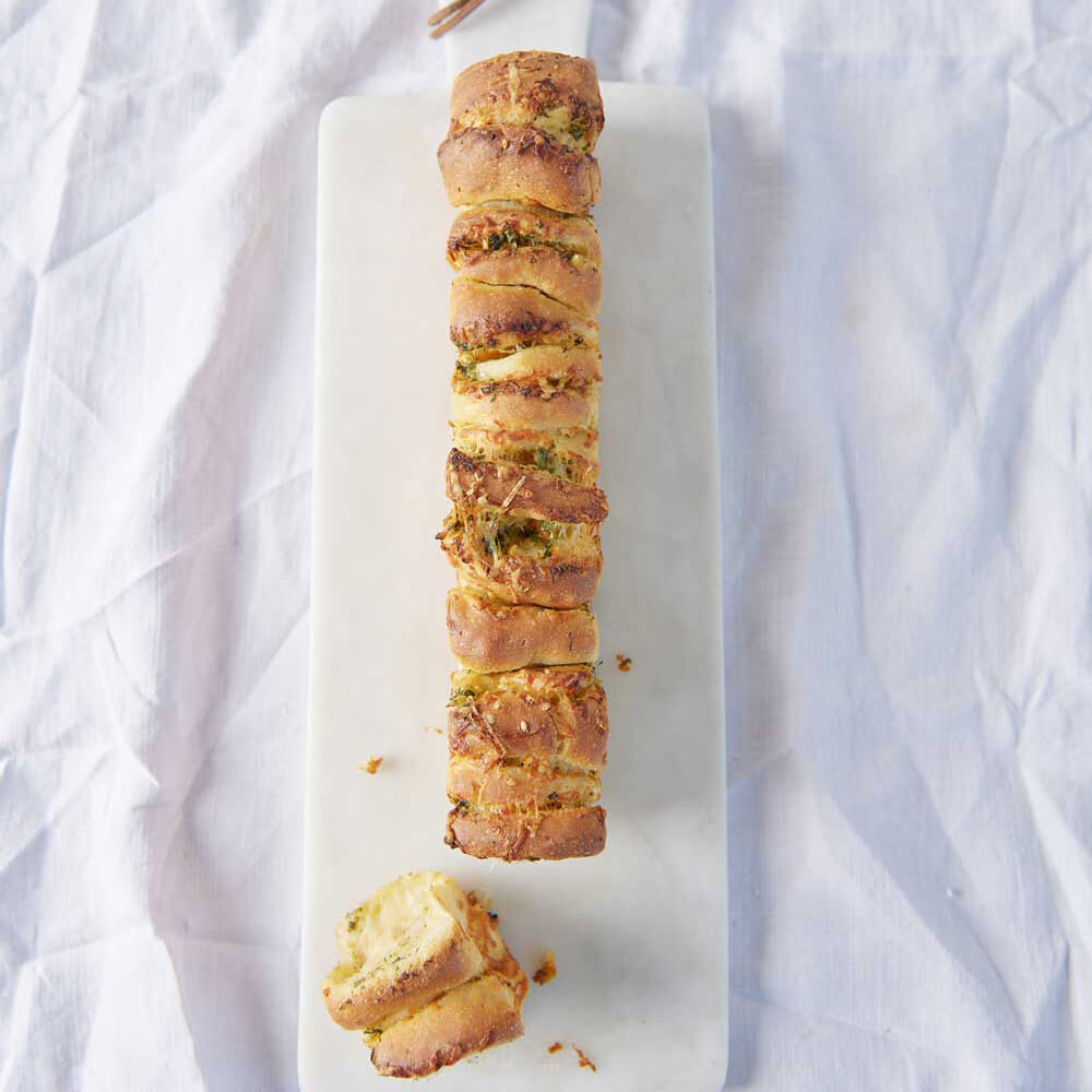 Zupfbrot mit einer Partystangenform gebacken, mit Käse überbacken auf einem weißen Brett, welches auf einem weißen Tuch steht.