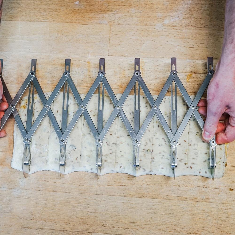 Es wird gezeigt wie der Teigschneider an einem Teig verwendet wird. Der Teig liegt auf einer braunen Arbeitsfläche.