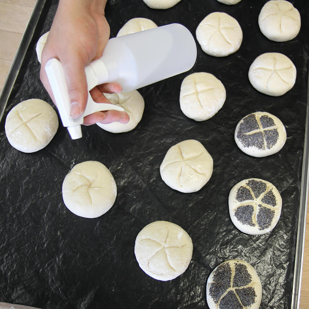 Mit dem Feinsprüher werden Brötchen befeuchet, die auf einem mit Dauerbackfolie belegtem Blech liegen. Die Brötchen auf der rechten Seite sind schon mit Saaten bedeckt. Die linken werden gerade besprüht um sie danach in Saaten zu tauchen