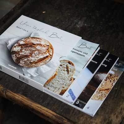 Backbuch-Set: Unser täglich Brot Band 1-4 Backbuchset. Die Bücher sind aufgefächert und schauen aus dem Schuber heraus. Der Hintergrund ist braun-schwarz