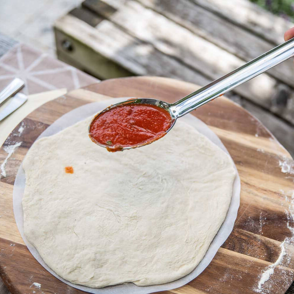 Ausgezogener Teig für Pizza auf rundem Backpapier auf einer Holzarbeitsplatte. Mit einem Löffel wird Tomatensoße darauf verteilt.