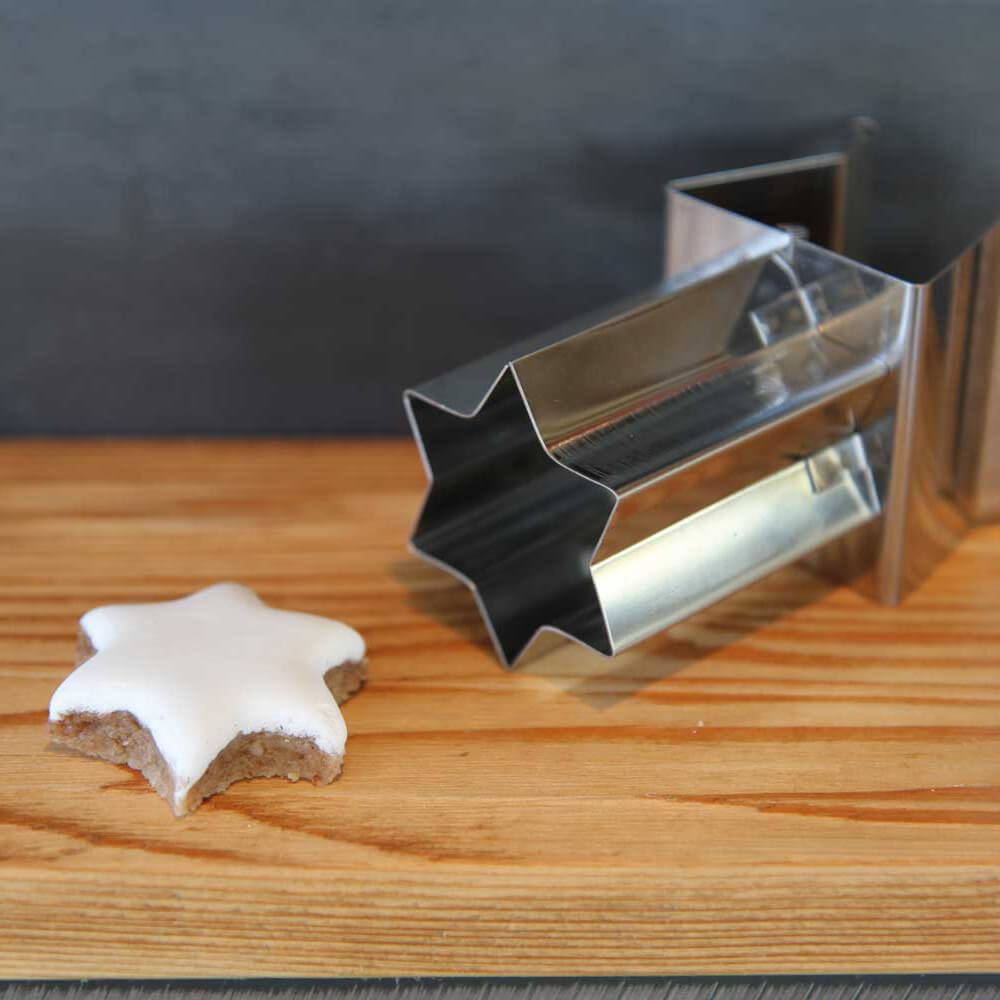 Links liegt ein Zimtstern mit weißer Glasur und rechts davon liegt ein Zimtsternausstecher. Beides liegt auf einer hellbraunen Oberfläche