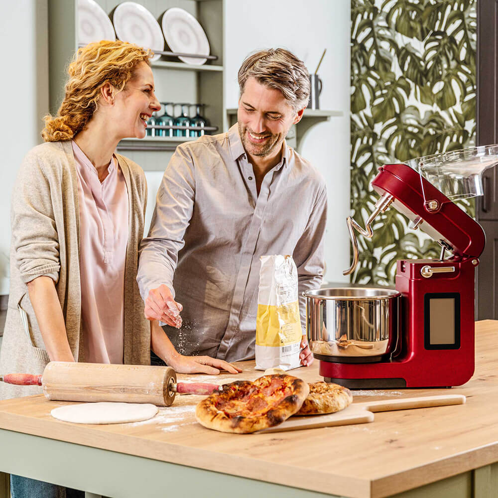 NOVA Teigknetmaschine in der Farbe Rot auf einer Küchenarbeitsplatte. Daneben liegen zwei frisch gebackene Pizzen und ein Mann und eine Frau schauen die Teigknetmaschine an.