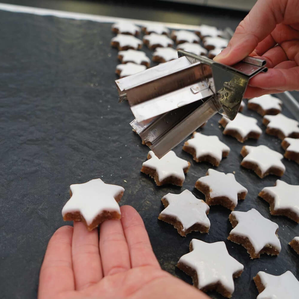 Jemand legt einen fertigen Zimtstern mit Glasur auf eine schwarze Dauerbackfolie und hält in der rechten Hand einen Zimtsternausstecher