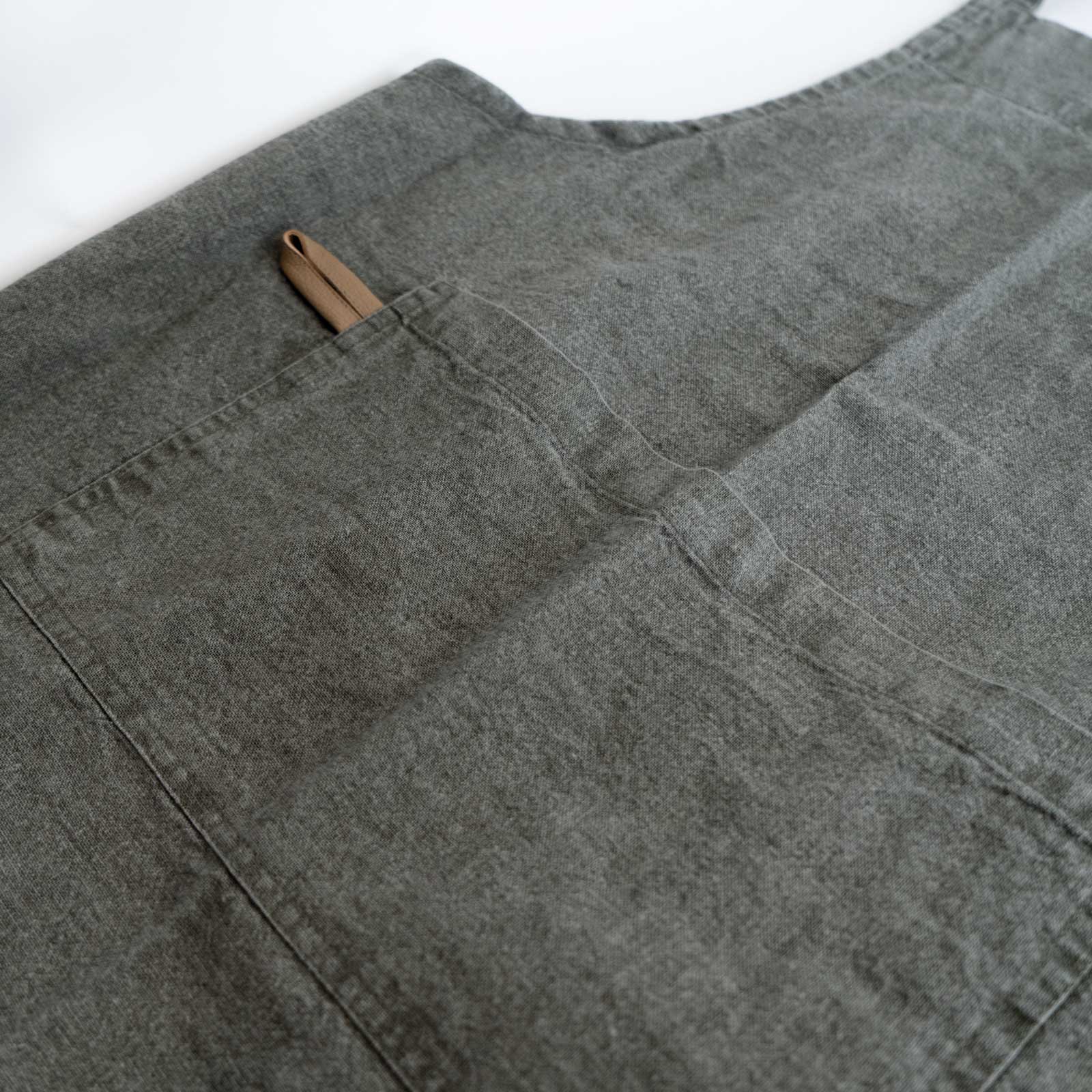 Schürze "MYRNA" oliv Auf dem Bild befindet sich ein Ausschnitt einer oliven Schürze auf dem zu erkennen ist, das die schürze eine Tasche besitzt.