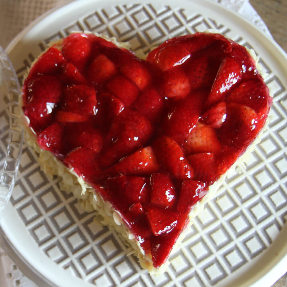 Kuchen in Herzform mit einer Vanillecreme und frischen Erdbeeren oben drauf, auf einer weiß-grau karierten Kuchenplatte serviert. Am Rand ist der Kuchen mit Mandelblättchen verziert.