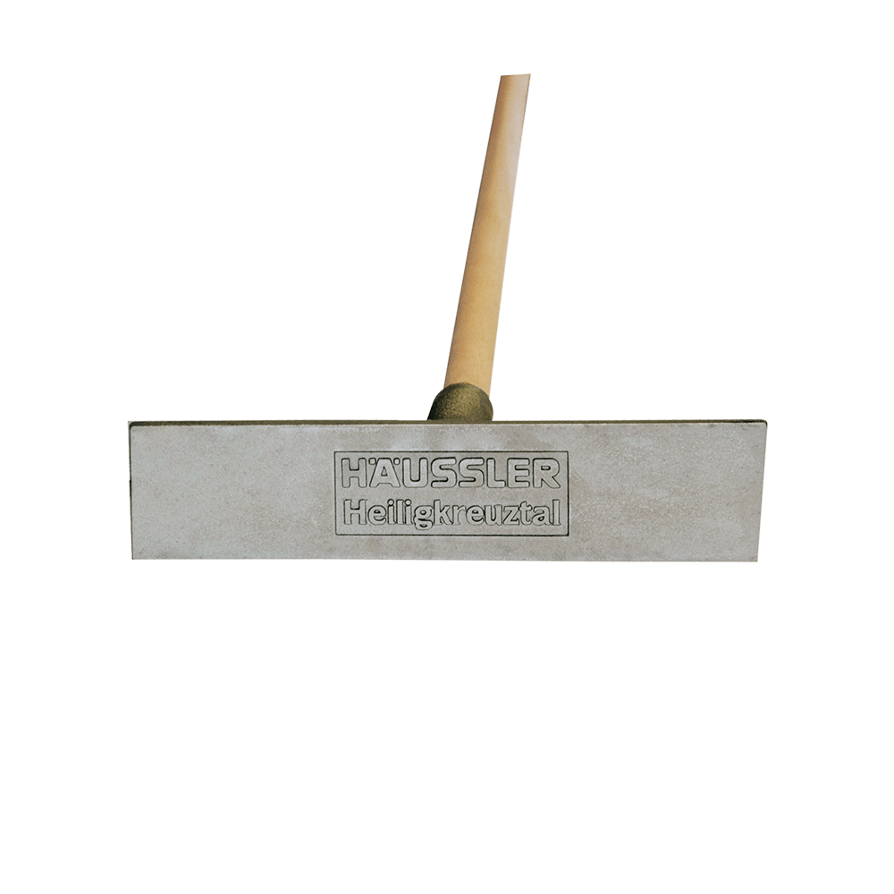 Ascheausziehkrücke Recheckige, aus Aluminiumgussgefertigte Ascheausziehkrücke mit der Prägung "Häussler Heiligkreuztal". Der Stiel ist aus hellem Holz gefertigt.