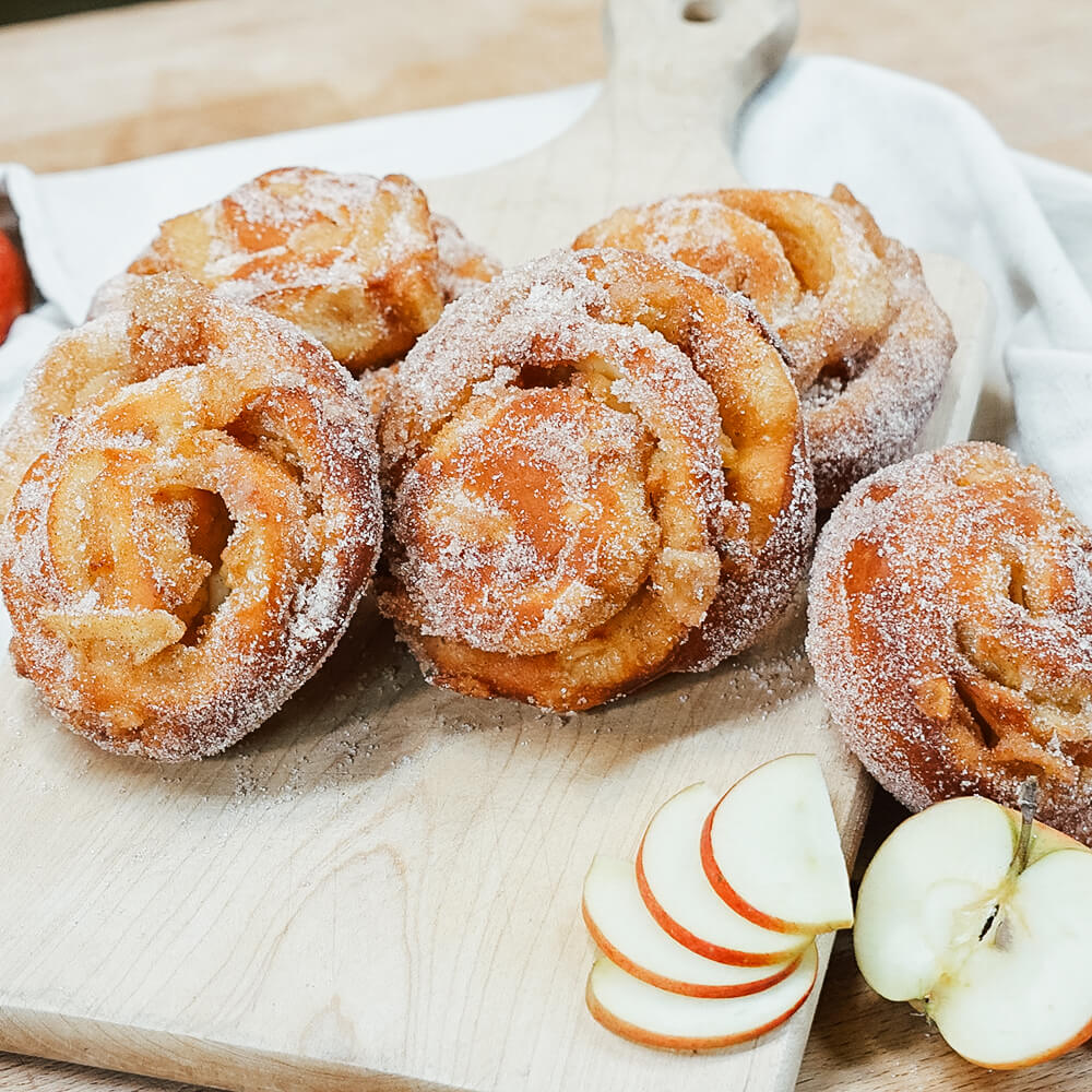 Rezept für Apfelkrapfen mehrere Apfelkrapfen gewälzt in einer Zimt-Zucker Mischung, serviert auf einem Backschieber mit eine paar frischen Apfelscheiben