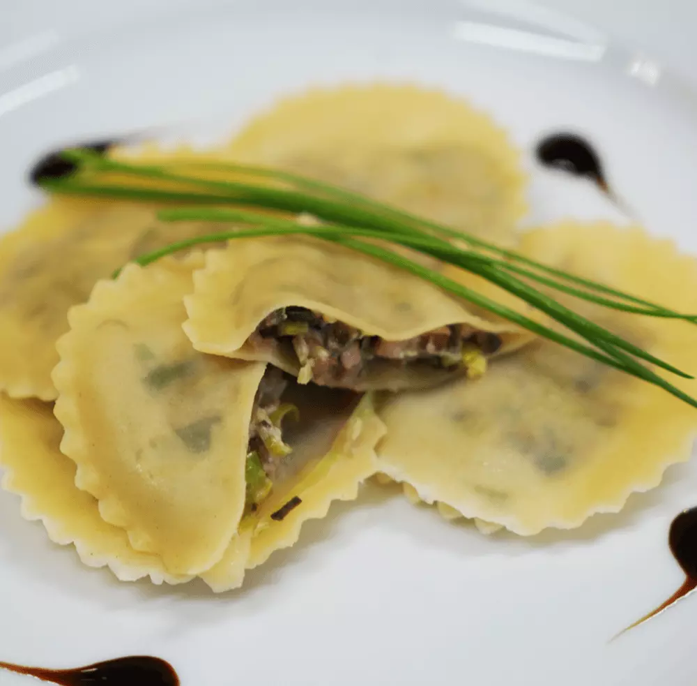 mehrere Ravioli, die auf einem Teller liegen, von denen zwei Ravioli angeschnitten sind. Auf den Ravioli liegt etwas Schnittlauch. Der Teller ist dekoriert mit mehreren Tropfen einer Soße.