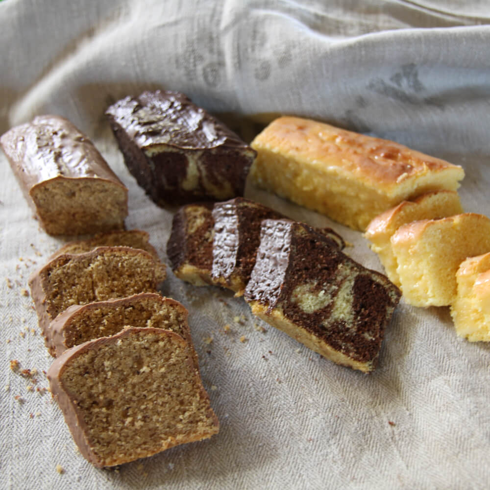 Rezept für Rührkuchen Variationen 3 Rührkuchen Varationen in Kastenform, zur Hälfte in Stücke geschnitten. Ein Nusskuchen, ein Marmorkuchen und ein normaler Rührkuchen auf einem grauen Tuch