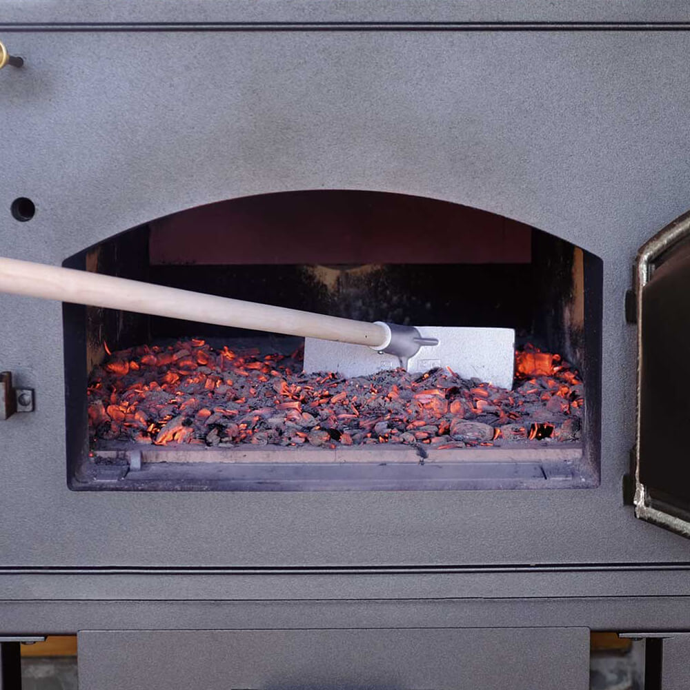 Ascheausziehkrücke Holzbackofen mit geöffneter Tür. Darin ist Glut verteilt. Diese wird mit Hilfe einer Ascheausziehkrücke verteilt.