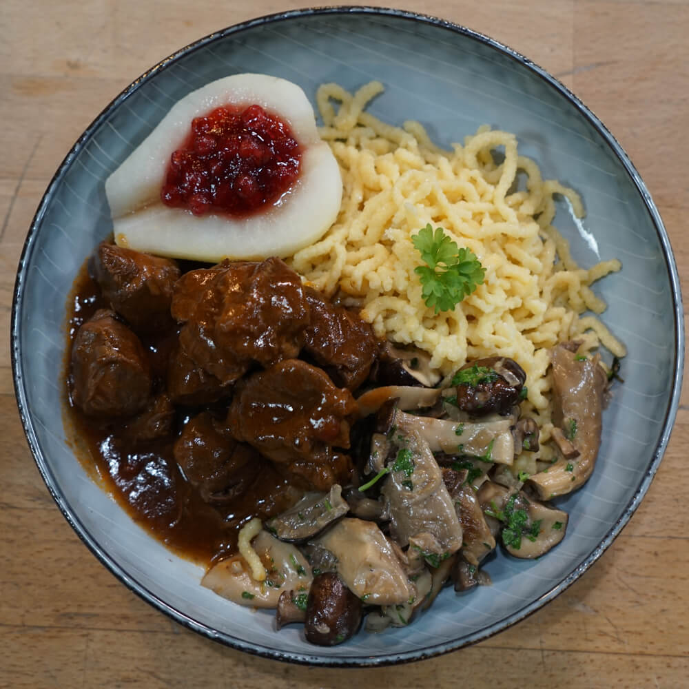Rezept für Wildgulasch Menü selbstgemachtes Wildgulasch in dunkler Soße, mit frischen Spätzel und einem leckeren Pilzragout mit frischen Käutern und einer halben Birne mit Preiselbeeren auf einem Blauen Teller serviert