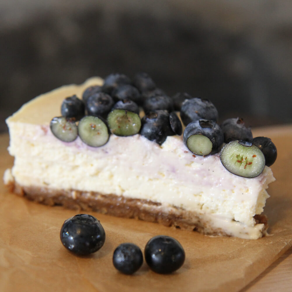 Rezept für New York Cheesecake 1 Stück cremiger Cheesecake mit Blaubeeren oben drauf auf einer Holzplatte serviert