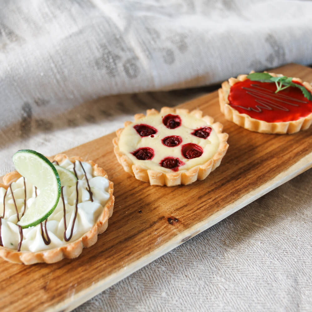 knusprige Tartlettes mit Cheescake Füllung, Himbeerfüllung und Limetten-Mascarpone Füllung auf einem Holzbrett serviert