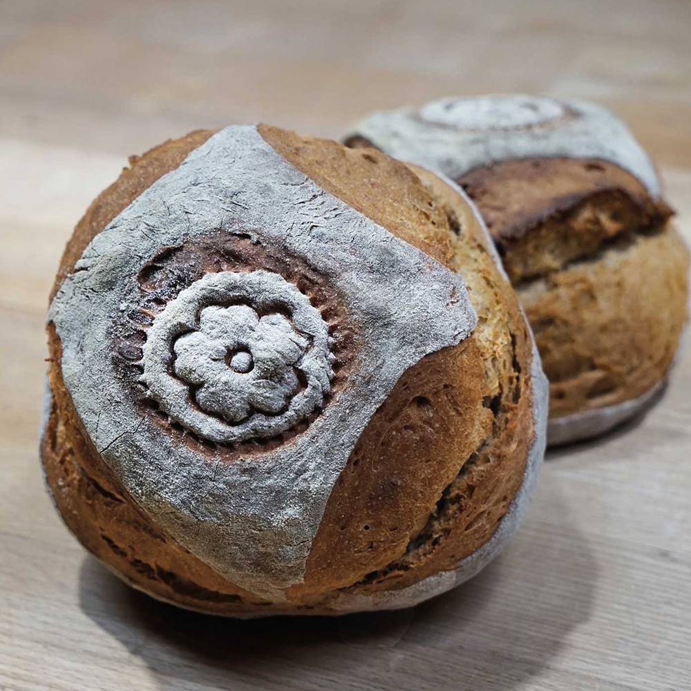 Brotstempel mit Bildmotiv Blume 2 Brotlaibe liegen nebeneinander auf einem Holztisch. Der vordere Laib ziert ein Blumen-Motiv in der Mitte