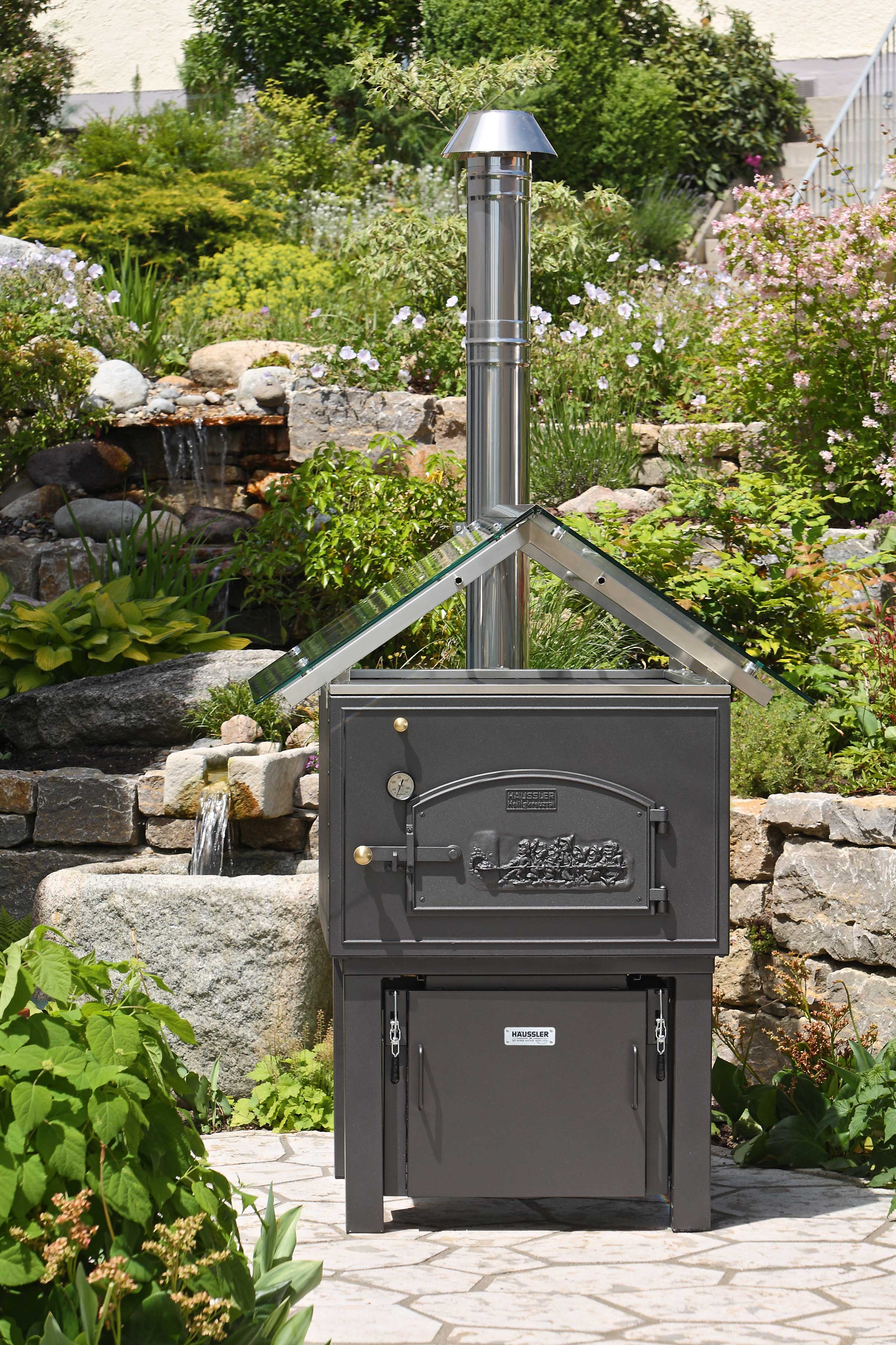 Holzbackofen mit einer Brennerkassette auf einer Terrasse. Im HIntergrund ist ein Steingarten mit viel Grün zu sehen