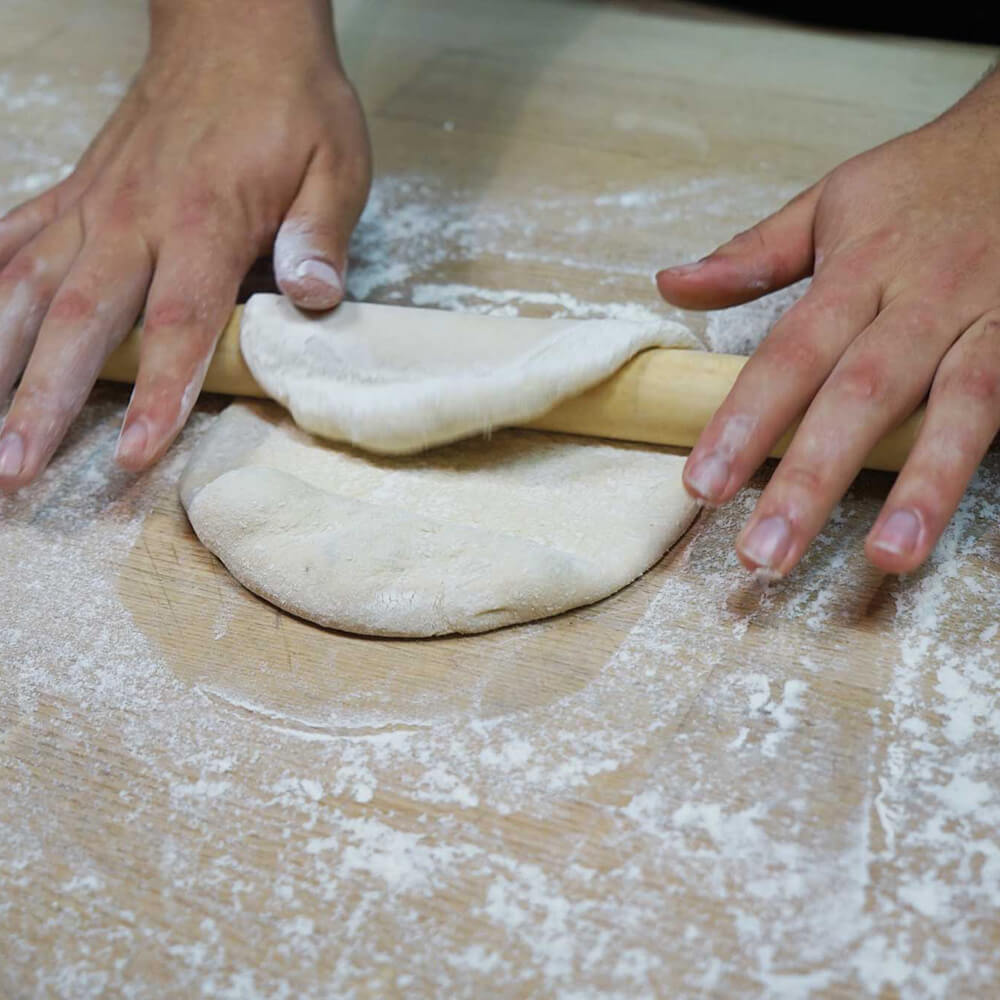 Rollholz aus Buche in der Anwendung beim Teigausrollen auf einer bemehlten Arbeitsfläche. Der Teig ist über das Rollholz geklappt.