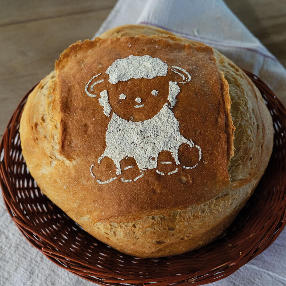 Ein Brotlaib liegt in einem geflochtenen Korb, der auf einem Tuch steht. Auf dem Brot ist ein Bild aus Mehl zu sehen in Form eines Schafs.