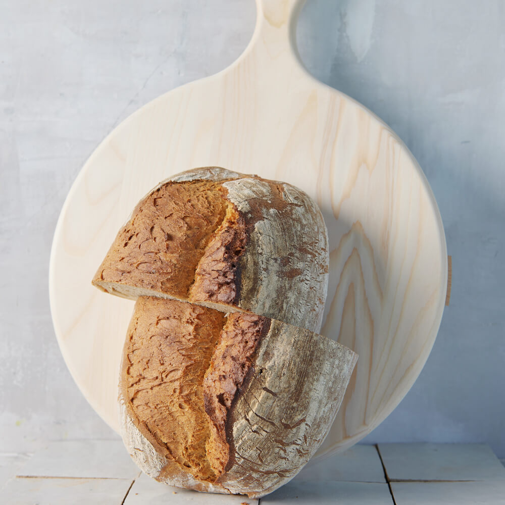 Rezept für Adelindes Schwäbisches Dinkelbrot längliches Schwäbisches Dinkelbrot, welches in der Mitte durchgeschnitten ist,aufgestellt vor einem runden Backbrett mit Griff, im Hintergrund ist eine hellgraue Wand