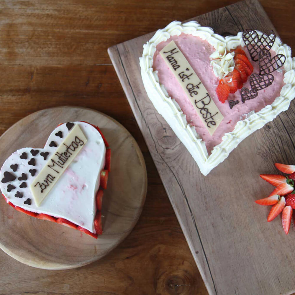 Es sind zwei Kuchen in Herzform zu sehen. Ein Kuchen ist mit weißer Glasur überzogen und es steht Zum Muttertag auf dem Kuchen. Der andere Kuchen hat eine pinke Glasur und die Aufschrift Mama ist die Beste