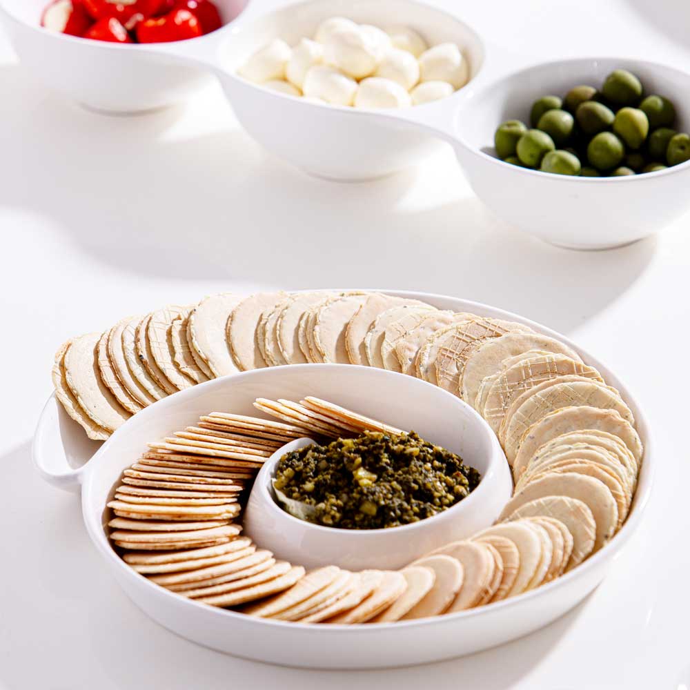 Servierteller Spirale, Farbe Weiß gefüllt mit runden Keksen und in der Mitte ist ein grüner Dipp, im HIntergrund sieht man 3 Servierschüsseln gefüllt mit kleinen Tomaten, Mozzarella und Oliven