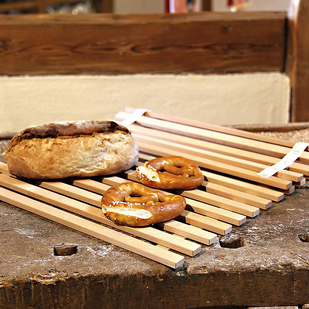 Auf dem Bild befindet sich ein Rollrost. Auf diesem liegen zwei Brezeln und ein schwäbisches Bauernbrot. ZUm liegt der Rollrost auf einer dunkelbraunen Oberfläche.