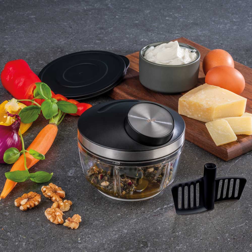 Eine Karotte, eine Zwiebel, eine Paprika und Walnüsse liegen auf einer schwarzen Küchenplatte. Daneben liegt ein Brett auf dem zwei Eier, Ein Stück Käse und Dip stehen. In dem Zerkleinerer der in der Mitte steht sind gehäckselte Kräuter und Walnusskerne